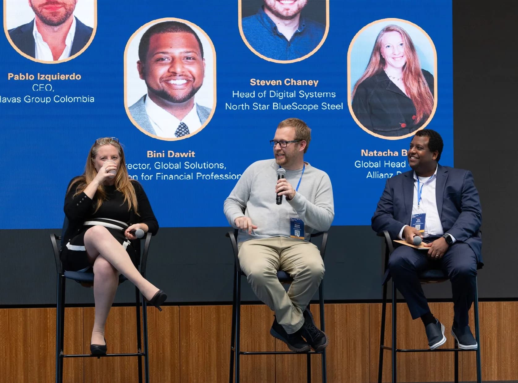 Steven Chaney speaking into a handheld microphone, seated among the other panellists with the session slide displayed behind them