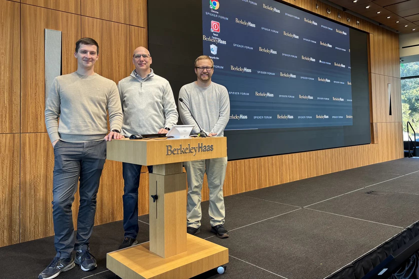 Three men standing side by side at the lectern on the Spieker Forum stage, the summit holding slide on the screen behind them and the seats still empty